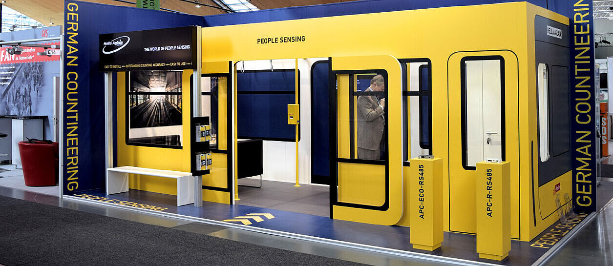 Hella Aglaia Messestand mit einer Nachbildung einer U-Bahn, einschließlich Sitzgelegenheiten, Griffstangen und Informationsdisplays. Oben hängt ein großes weißes Banner mit dem Firmenlogo und den Worten „GERMAN COUNTEINEERING · PEOPLE SENSING“.