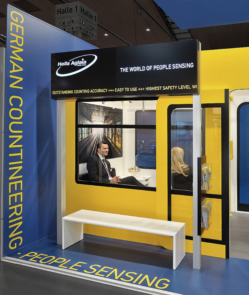 Messestand von Hella Aglaia in Form eines U-Bahn-Waggons mit gelb-blauer Gestaltung. Besucher unterhalten sich in einem nachgebildeten Abteil, während eine Person mit Aktenmappe vorbeigeht. Ein Bildschirm im Hintergrund zeigt Informationen zur Fahrgastzählungstechnologie.