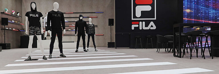 Mehrere Schaufensterpuppen tragen sportliche FILA-Outfits auf einem Messeboden, der wie ein Zebrastreifen gestaltet ist. Im Hintergrund sind beleuchtete Regale mit Schuhen, ein großes FILA-Logo sowie schwarze Tische und Stühle zu sehen.
