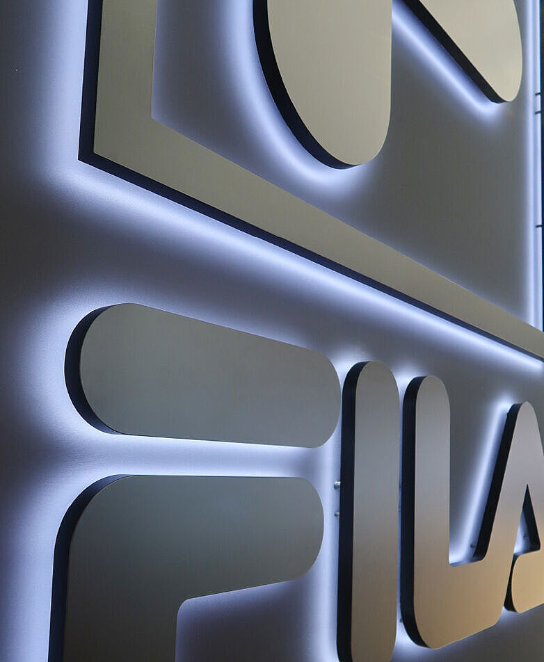 Ein großes, dreidimensionales FILA-Logo an einer dunklen Wand. Die Buchstaben sind erhaben und werden von einer dezenten weißen Hintergrundbeleuchtung umrahmt, wodurch ein eleganter Schwebeeffekt entsteht. Die Perspektive der Aufnahme ist leicht schräg, wodurch die Tiefe des Logos betont wird. Im Hintergrund sind unscharf Messe- oder Geschäftsumgebungen zu erkennen.