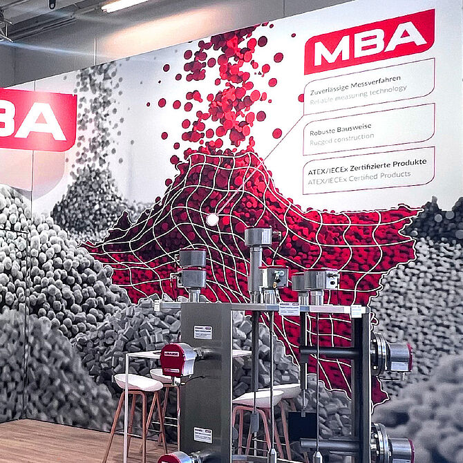 Ein Messestand von MBA Instruments mit einer auffälligen Wandgrafik aus roten und grauen Pellets, die durch ein weißes Gitternetz überlagert sind. Im Vordergrund sind technische Messgeräte mit mehreren Sensoren zu sehen. Im Hintergrund befinden sich eine Sitzgruppe mit weißen Barhockern und ein Stehtisch.