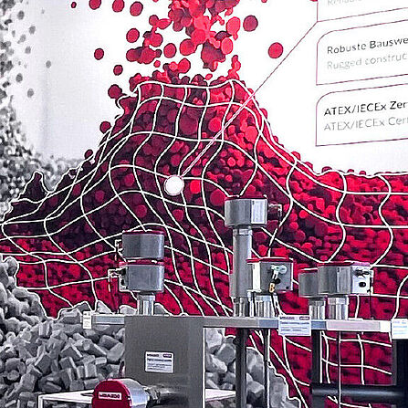 Ein Messestand von MBA Instruments mit einer auffälligen Wandgrafik aus roten und grauen Pellets, die durch ein weißes Gitternetz überlagert sind. Im Vordergrund sind technische Messgeräte mit mehreren Sensoren zu sehen. Im Hintergrund befinden sich eine Sitzgruppe mit weißen Barhockern und ein Stehtisch.