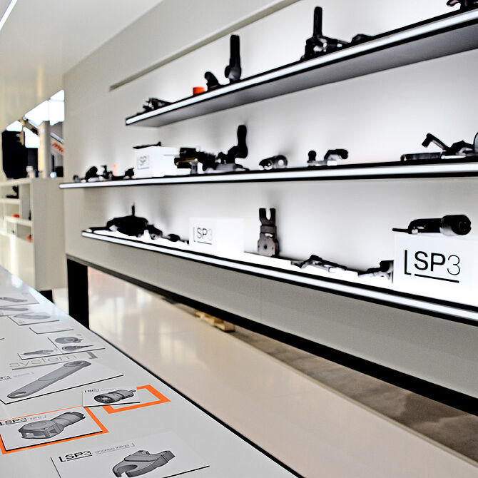 Ein stilvoll gestaltetes Ausstellungsregal mit verschiedenen schwarzen Bauteilen, die auf leuchtenden weißen Ablagen präsentiert werden. Auf einem Tisch davor liegen technische Zeichnungen und Produktbeschreibungen mit orangefarbenen Markierungen.