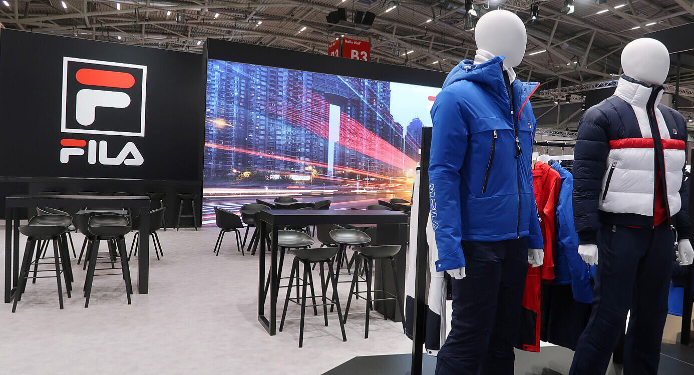 Ein FILA-Messestand mit zwei Schaufensterpuppen im Vordergrund, die moderne Winterjacken tragen – eine in Blau mit Kapuze, die andere in Blau, Weiß und Rot mit einer gesteppten Optik. Dahinter befindet sich eine Lounge-Ecke mit schwarzen Tischen und Barhockern. An der Wand ist ein großes FILA-Logo sowie eine LED-Wand mit einer Stadtaufnahme und roten Lichtspuren zu sehen. Im Hintergrund hängt weitere FILA-Bekleidung auf Kleiderständern. Der Stand hat eine moderne, elegante Atmosphäre mit schwarzem und neutralem Design.