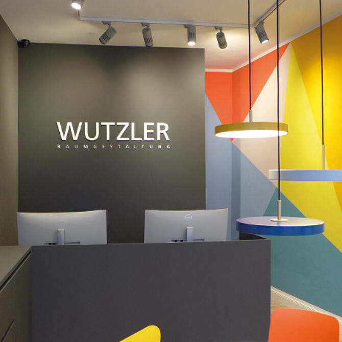 Ein modern gestaltetes Büro oder Empfangsbereich der Firma „Wutzler Raumgestaltung“ mit einer dunklen Akzentwand, bunten geometrischen Wandmustern und stilvollen Hängelampen. Zwei Arbeitsplätze mit Monitoren befinden sich im Vordergrund.