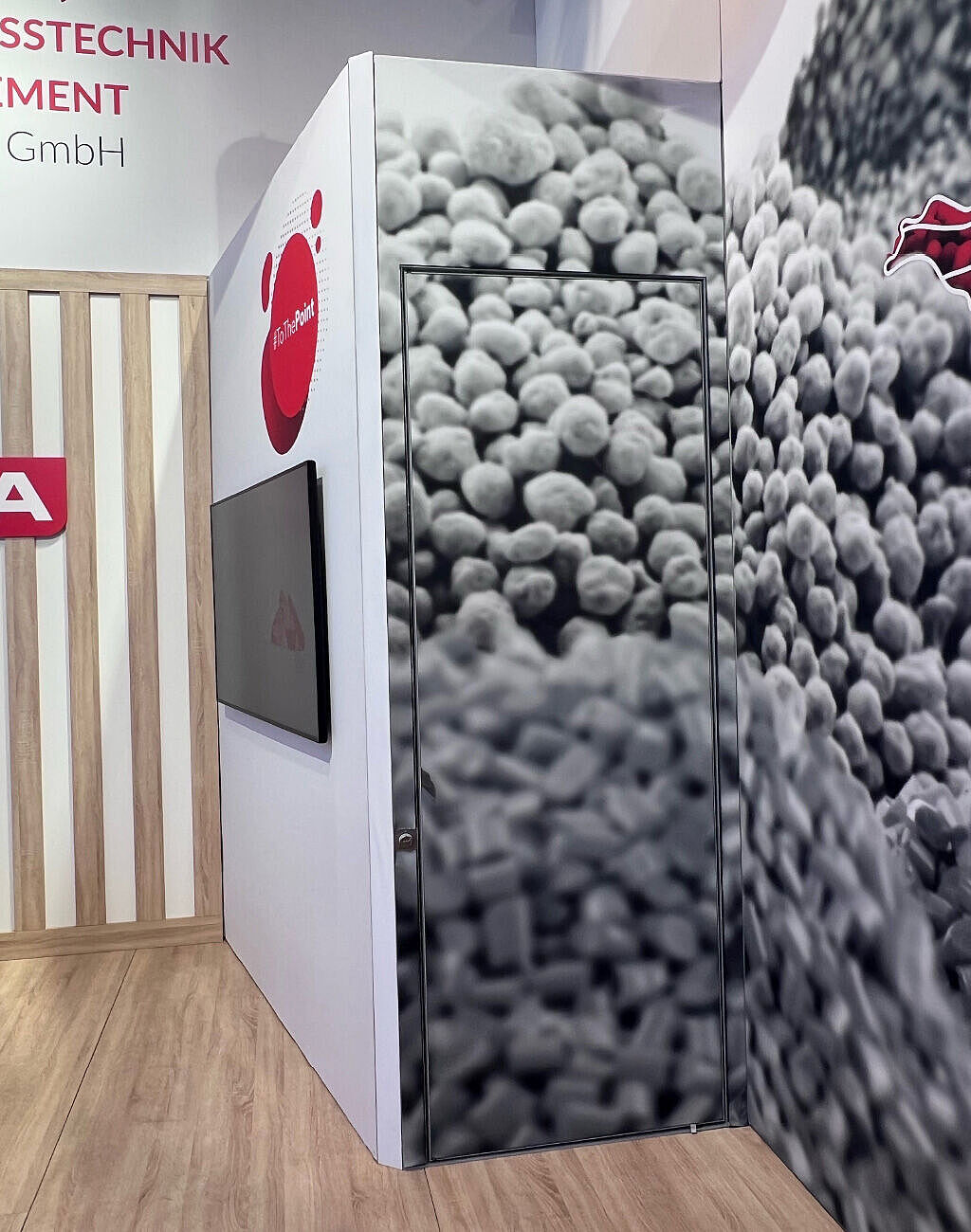 Eine geschickt getarnte Tür in einem Messestand, die mit einer großflächigen Grafik von Pellets bedruckt ist. Daneben befindet sich ein Bildschirm und eine Wand mit Holzlamellen. Der Boden hat eine Holzoptik.