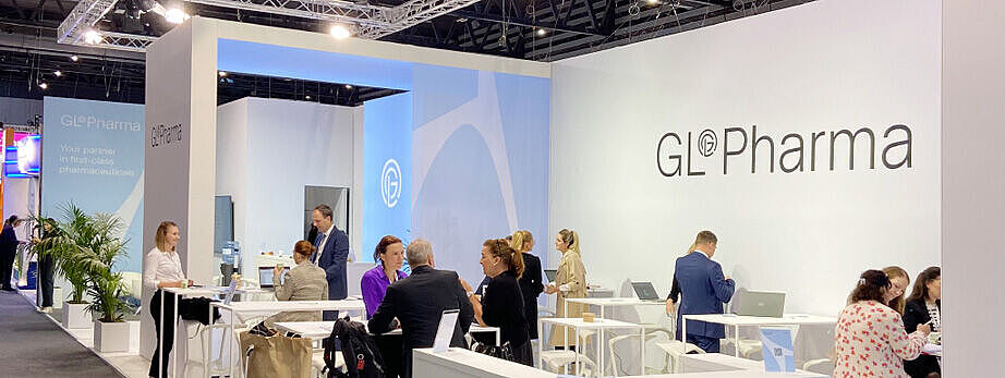 GL Pharma Messestand mit weißem Empfangstresen, Sitzgruppen und Geschäftsleuten in Gesprächen. Das Design ist hell und offen, mit weißen Möbeln und blauen Akzenten. Im Hintergrund Pflanzen und weitere Messestände.