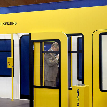 Messestand von Hella Aglaia in Gelb und Blau, gestaltet wie ein U-Bahn-Waggon. Ein Mann im Anzug steht im Hintergrund und nutzt sein Smartphone. Zwei gelbe Säulen mit der Aufschrift „APC-ECO-RS485“ und „APC-R-RS485“ stehen vor dem Stand, der mit Fenstern, Türen und Haltestangen eine Bahnumgebung simuliert.