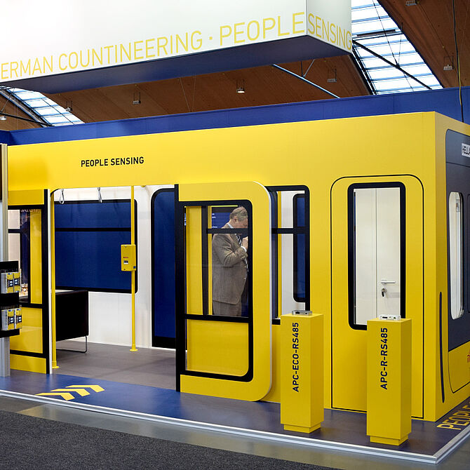 Messestand von Hella Aglaia in Gelb und Blau, gestaltet wie ein U-Bahn-Waggon. Ein Mann im Anzug steht im Hintergrund und nutzt sein Smartphone. Zwei gelbe Säulen mit der Aufschrift „APC-ECO-RS485“ und „APC-R-RS485“ stehen vor dem Stand, der mit Fenstern, Türen und Haltestangen eine Bahnumgebung simuliert.