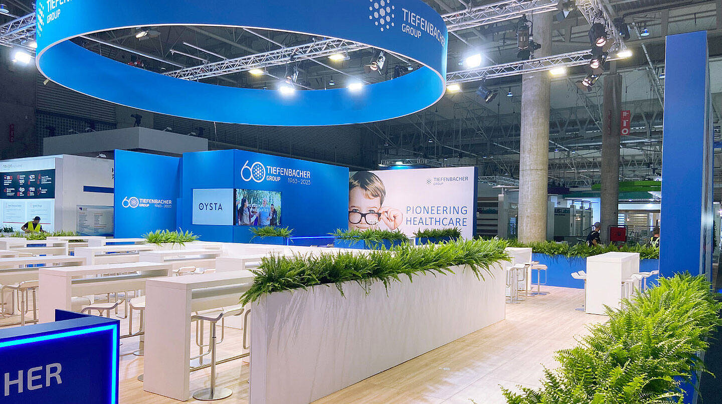 Ein moderner Messestand der Tiefenbacher Group mit blau-weißem Design und einer großen, leuchtenden Deckenkonstruktion. Helle Sitzbereiche mit weißen Tischen und Barhockern sind in einer offenen Raumgestaltung angeordnet. Zahlreiche grüne Pflanzen schmücken den Stand und sorgen für eine freundliche Atmosphäre. Auf großen Bildschirmen sind Informationen zur Tiefenbacher Group zu sehen. Im Hintergrund sind weitere Messestände und Besucher erkennbar.