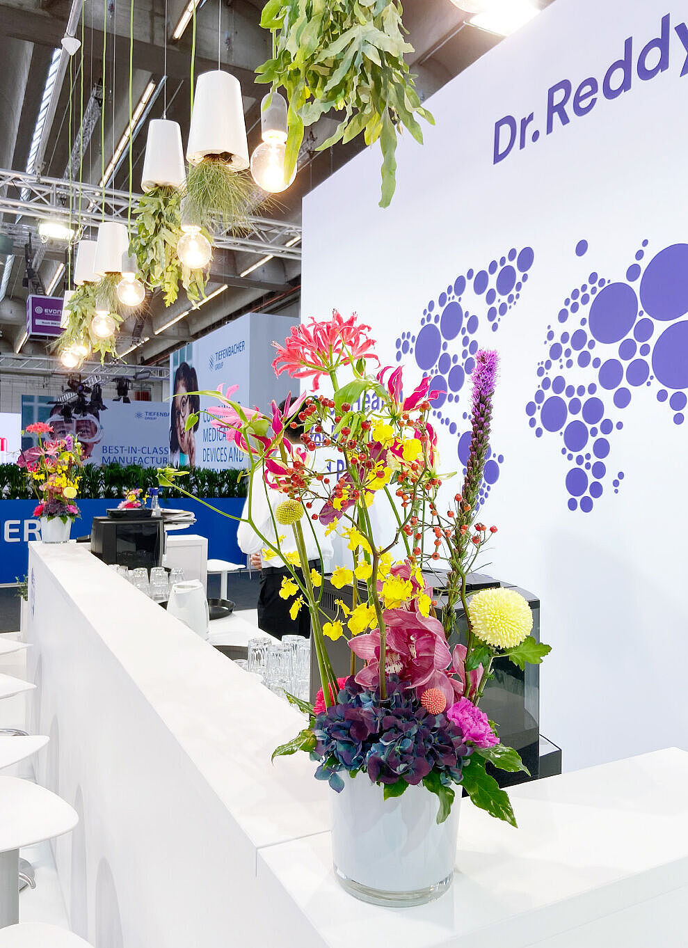 Ein eleganter Messestand mit einer weißen Theke, auf der sich bunte Blumenarrangements in weißen Vasen befinden. Darüber hängen moderne Lampen mit warmem Licht, kombiniert mit umgedrehten weißen Blumentöpfen mit herabhängenden Pflanzen. Im Hintergrund eine große Wand mit dem Logo von Dr. Reddy’s und einer stilisierten Weltkarte aus violetten Kreisen. Ein Barista oder Standmitarbeiter in weißem Hemd steht hinter der Theke.
