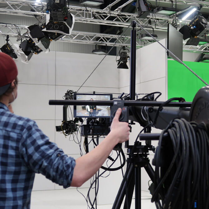Ein Kameramann mit roter Cap und kariertem Hemd bedient eine professionelle Kamera auf einem Stativ in einem Hightech-Studio. Vor ihm zeigt ein Kontrollmonitor das laufende Live-Bild. Im Hintergrund sind mehrere Studiolichter an Traversen montiert, und ein Greenscreen ist sichtbar. Die step one GmbH setzt modernste Technik für digitale und hybride Events ein.
