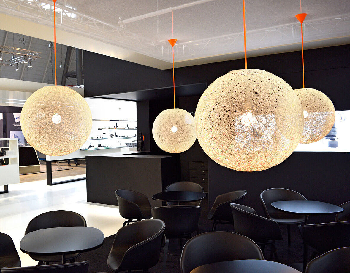 Elegante Lounge auf einem Messestand mit schwarzen Stühlen und Tischen. Große kugelförmige Lampen aus feinem Drahtgeflecht hängen von der Decke an orangefarbenen Kabeln. Im Hintergrund sind beleuchtete Produktregale und eine moderne Theke zu sehen.