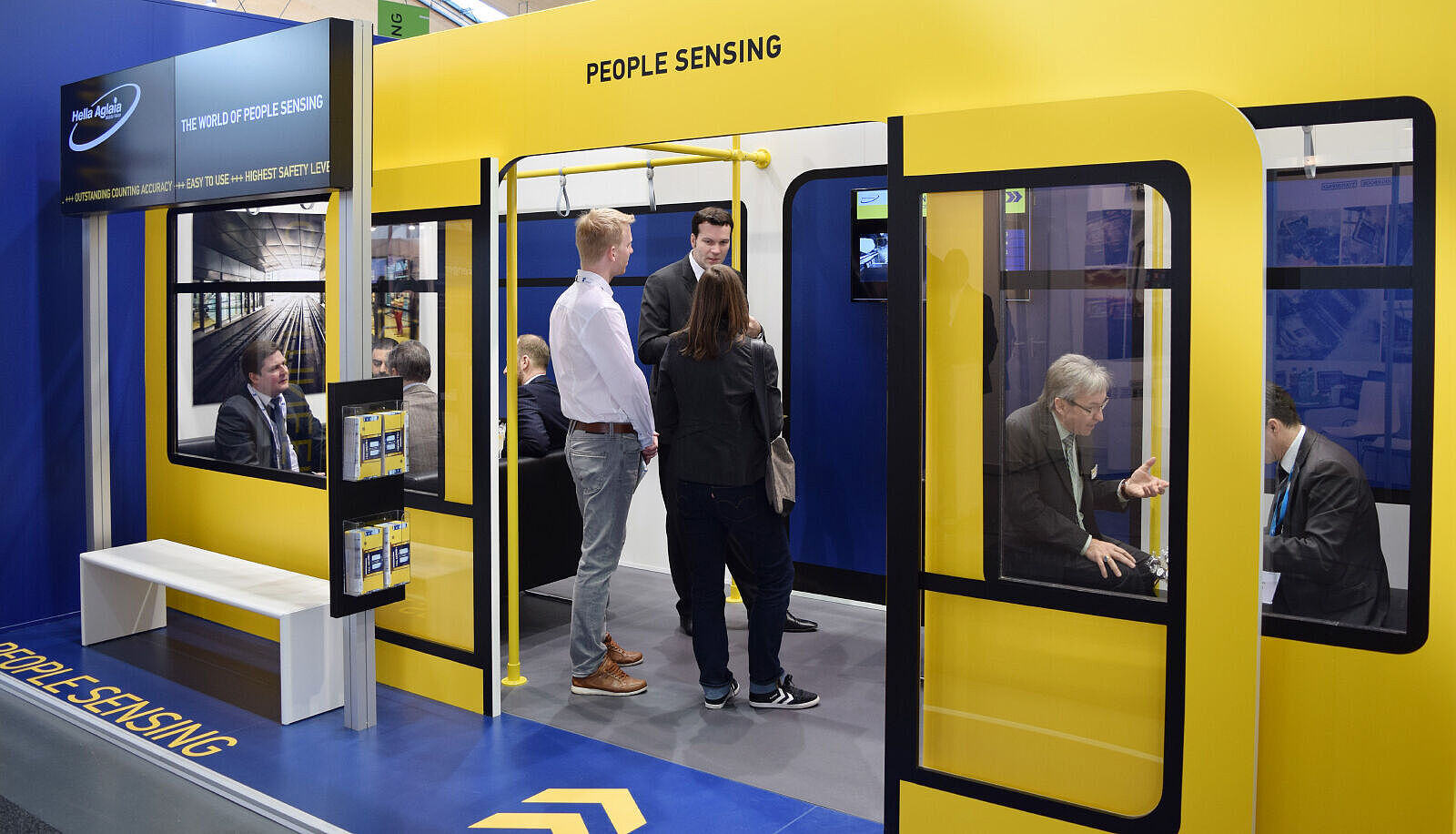 Messestand von Hella Aglaia mit einer gelb-schwarzen U-Bahn-Kulisse. Menschen stehen und sitzen im Nachbau eines Zugabteils, während Gespräche über Technologien zur Personenerfassung geführt werden.