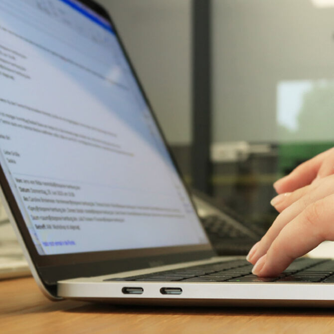 Nahaufnahme eines Laptops mit geöffneter E-Mail-Anwendung, während eine Person auf der Tastatur tippt. Im Hintergrund ein Büro mit Glaswand.