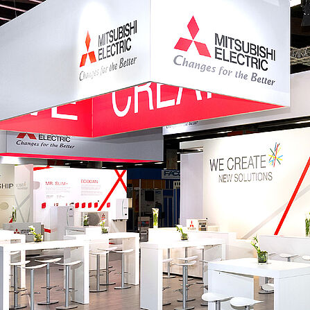 Ein moderner Mitsubishi Electric Messestand mit weiß-rotem Design und großen Deckenbannern mit dem Firmenlogo. Der Stand bietet Informationsbereiche zu Klimasystemen, energieeffizienten Lösungen und neue Technologien. Weiße Sitzgelegenheiten mit Barhockern und grünen Pflanzen schaffen eine angenehme Atmosphäre für Gespräche.