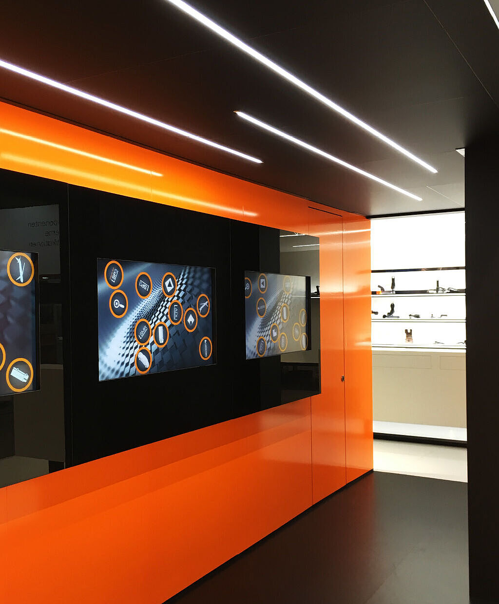 Ein interaktives digitales Display mit mehreren Bildschirmen, die eine grafische Benutzeroberfläche mit Symbolen zeigen. Der Raum hat eine moderne, schwarz-orange Farbgestaltung mit LED-Streifen an der Decke und reflektierenden Oberflächen.
