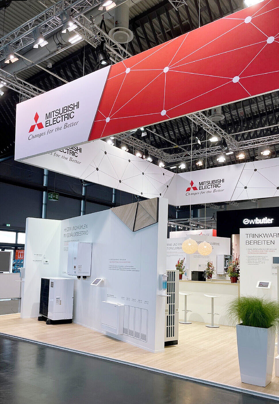 Ein eleganter Mitsubishi Electric Messestand mit weiß-rotem Design und modernen Heiz- und Kühlsystemen. Die Wände zeigen Infografiken und Geräte zur energieeffizienten Klimatisierung und Warmwasserbereitung. Eine stilvolle Bar mit Blumen und Sitzgelegenheiten lädt Besucher zum Verweilen ein.
