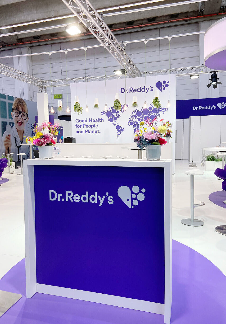 Ein einladender Messestand von Dr. Reddy’s mit einem Empfangstresen in Lila und Weiß, auf dem das Firmenlogo zu sehen ist. Im Hintergrund eine große Wand mit einer stilisierten Weltkarte aus lila Kreisen und dem Motto „Good Health for People and Planet.“ Mehrere weiße Hängeleuchten mit grünen Pflanzen hängen von der Decke. Der Stand verfügt über weiße Sitzmöbel, moderne Stehtische und Dekorationen mit bunten Blumen. Eine freundliche Atmosphäre wird durch helles Licht und ein klares, professionelles Design geschaffen.