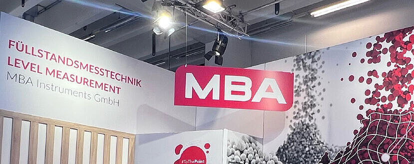 Ein Messestand von MBA Instruments GmbH mit großem Wanddesign aus rot-weißen Partikeln, einem Infobereich mit Monitor, Stehtischen und ausgestellten Messgeräten. Die Beleuchtung ist professionell installiert, und verschiedene Infotafeln heben die Eigenschaften der Produkte hervor.