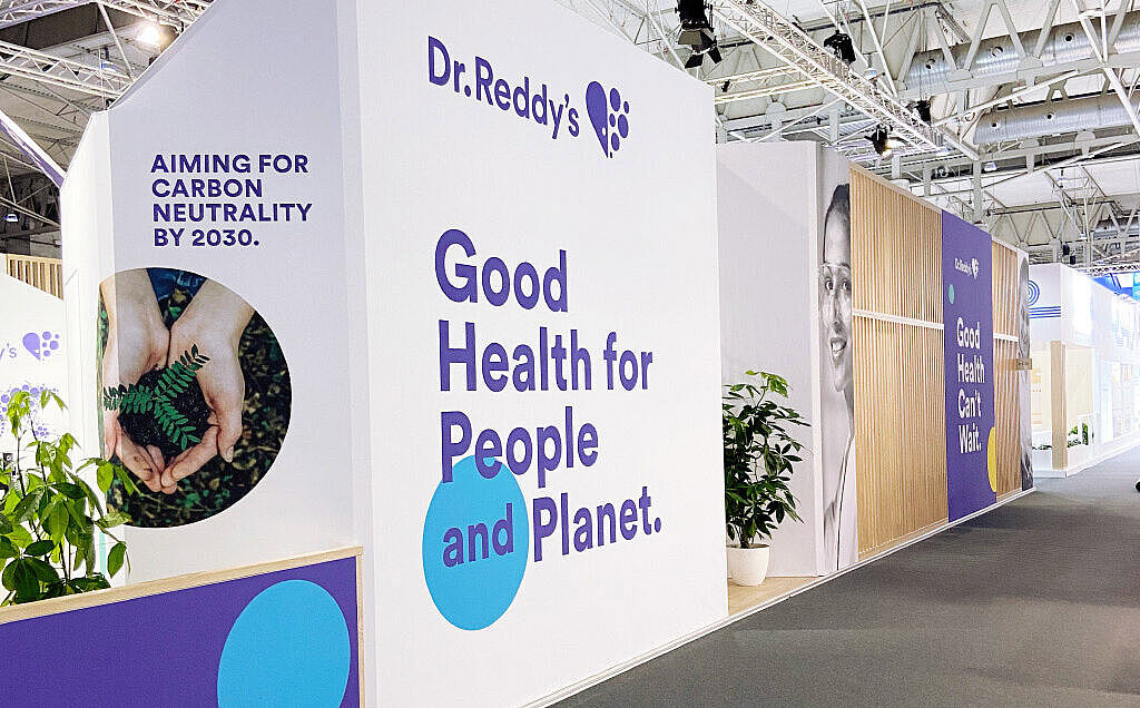 Ein moderner Messestand von Dr. Reddy’s mit einer weißen Wand, auf der in großen blauen Buchstaben „Good Health for People and Planet.“ steht. Links ist ein rundes Bild von Händen, die eine kleine Pflanze halten, mit dem Text „Aiming for carbon neutrality by 2030.“ Die Standgestaltung enthält blaue und gelbe Akzente sowie grüne Pflanzen in weißen Töpfen. Der Boden ist mit dunklem Teppich ausgelegt, und über dem Stand sind Messebeleuchtungen installiert.