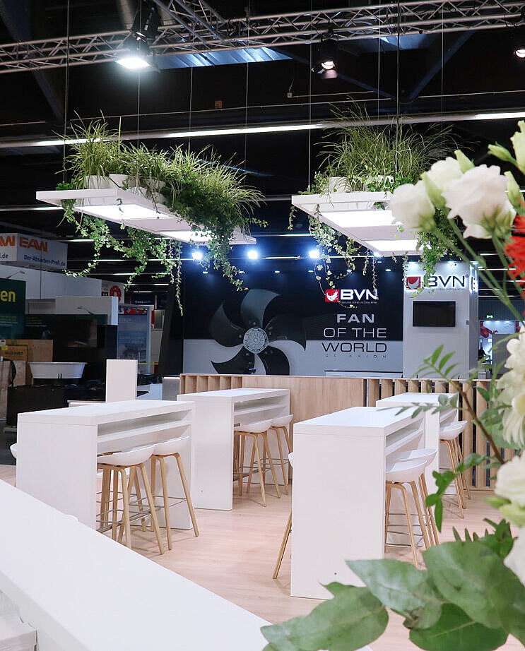 Ein stilvoll gestalteter Messestand mit weißen Stehtischen und Barhockern aus Holz. Im Vordergrund ein großer Strauß mit weißen und roten Blumen. Über dem Stand hängen rechteckige Pflanzkästen mit grünen, herabhängenden Pflanzen. Im Hintergrund ist eine dunkle Messewand mit dem Logo von BVN und der Aufschrift 'FAN OF THE WORLD' zu sehen. Weitere Messestände und Besucher sind in der Ferne erkennbar.