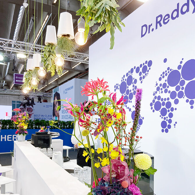 Eine Nahaufnahme des Dr. Reddy’s Messestandes zeigt eine weiße Theke mit bunten Blumenarrangements in Vasen. Im Hintergrund ist eine Wand mit dem Firmenlogo und einer Weltkarte aus violetten Kreisen zu sehen. Über der Theke hängen Lampen mit umgedrehten weißen Blumentöpfen, aus denen grüne Pflanzen wachsen. Im hinteren Bereich des Bildes sind weitere Stände mit Informationsmaterial und Werbetafeln sichtbar.