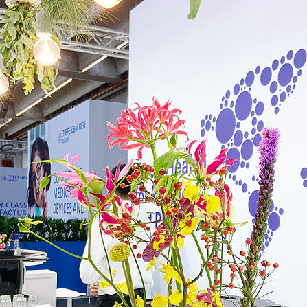 Eine Nahaufnahme des Dr. Reddy’s Messestandes zeigt eine weiße Theke mit bunten Blumenarrangements in Vasen. Im Hintergrund ist eine Wand mit dem Firmenlogo und einer Weltkarte aus violetten Kreisen zu sehen. Über der Theke hängen Lampen mit umgedrehten weißen Blumentöpfen, aus denen grüne Pflanzen wachsen. Im hinteren Bereich des Bildes sind weitere Stände mit Informationsmaterial und Werbetafeln sichtbar.