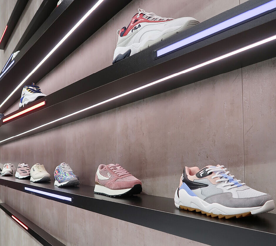 Ein Wandregal mit schräg angebrachten, schwarzen Ablagen, auf denen verschiedene Sneaker-Modelle ausgestellt sind. Die Regale sind mit schmalen LED-Lichtstreifen in Weiß, Rot und Blau beleuchtet. Der Hintergrund besteht aus einer Betonoptik-Wand, die den modernen und minimalistischen Stil der Präsentation unterstreicht. Der Fokus liegt auf einem grauen, pink und blauen Sneaker im Vordergrund, während weitere Schuhe weiter hinten entlang der Regalreihe sichtbar sind.