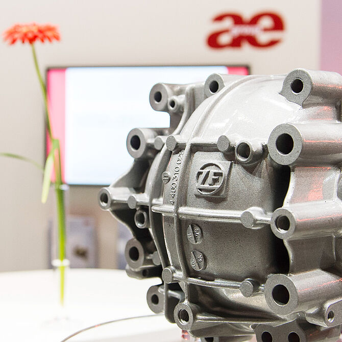 Nahaufnahme eines silbernen Metallbauteils. Das Bauteil ist ausgestellt auf einer Messe, im Hintergrund sind ein Bildschirm mit Präsentation, eine rote Blume und ein Firmenlogo sichtbar.