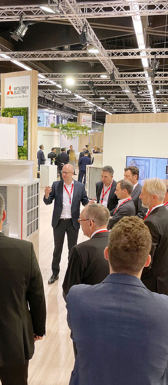 Ein Mann in einem dunklen Anzug mit rotem Messeausweis erklärt einer Gruppe von Geschäftsleuten eine Klimaanlagen-Einheit auf einem Messestand von Mitsubishi Electric. Die Teilnehmer, überwiegend in Anzügen und mit roten Namensschildern, hören aufmerksam zu. Der Stand ist modern gestaltet, mit Holzelementen, grünen Pflanzen und leuchtenden Displays. Im Hintergrund sind weitere Messebesucher und Stände zu sehen.