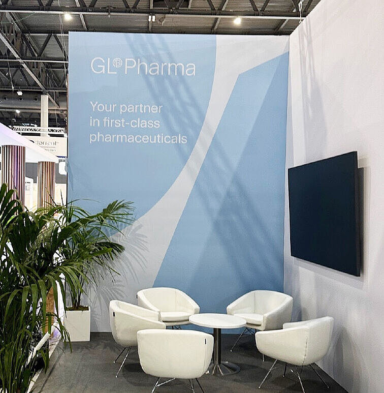 Messestand von GL Pharma mit vier weißen Sesseln, zwei runden Tischen und einem großen blauen Werbebanner mit Firmenlogo und Slogan. An der Wand hängt ein schwarzer Bildschirm. Links stehen große grüne Pflanzen. Der Boden ist teils weiß, teils dunkelgrau.