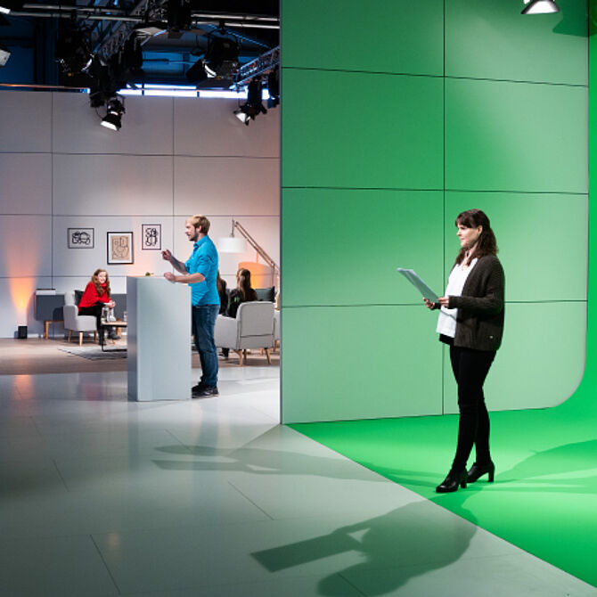 In einem modernen Studio stehen eine Frau mit Notizen vor einem Greenscreen und ein Mann an einem weißen Podium. Im Hintergrund befindet sich eine stilvolle, beleuchtete Gesprächsrunde mit mehreren Personen auf Sofas. Über ihnen sind Studiolichter angebracht, die für eine professionelle Atmosphäre sorgen. Die step one GmbH setzt innovative Technik für hybride Events ein.