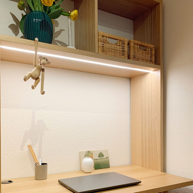 Ein stilvoll eingerichteter Holz-Schreibtisch mit eingebautem Regal, indirekter Beleuchtung und dekorativen Elementen wie einer Vase mit gelben Blumen, einem Holzaffen und geflochtenen Körben. Ein Laptop und Schreibutensilien runden das gemütliche Arbeitsambiente ab.