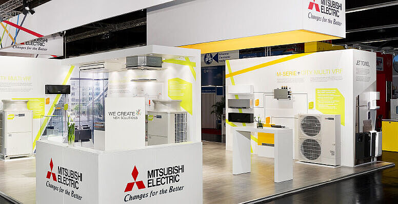 Ein heller, moderner Messestand von Mitsubishi Electric mit weißen und gelben Designelementen. Verschiedene Klimasysteme, darunter VRF- und M-Serie-Modelle, sind ausgestellt. Infotafeln erläutern technische Details, während digitale Bildschirme und Pflanzen für eine einladende Atmosphäre sorgen.