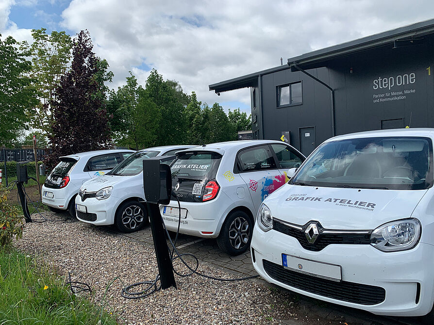 Vier weiße Elektroautos der step one GmbH an Ladestationen vor einem modernen Firmengebäude mit Firmenlogo von der step one GmbH.