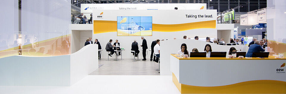 Ein offener und modern gestalteter Messestand von EEW mit einem weißen und orangefarbenen Design. Vorne befindet sich ein großer Empfangstresen mit mehreren Mitarbeiterinnen. Im Hintergrund gibt es eine Lounge mit Sitzgelegenheiten, an denen Geschäftsleute Gespräche führen. Ein großer Bildschirm zeigt Informationen über das Unternehmen, und das Motto „Taking the lead.“ ist auf der Wand zu lesen. Die Messehalle ist mit zahlreichen Lichtern ausgestattet, die den Stand hell und einladend wirken lassen.