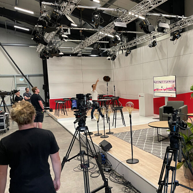 A live event setting with a digital component: a large screen shows a live broadcast while people interact. Lighting and stage effects create a dynamic atmosphere.