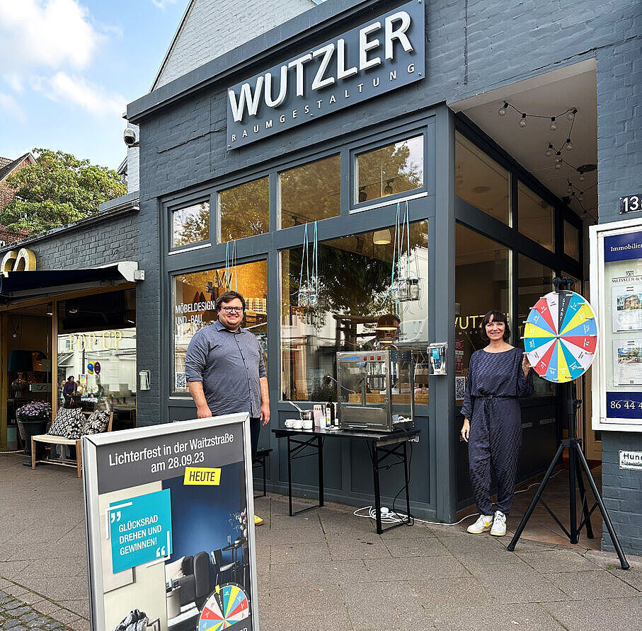Zwei Personen stehen vor einem Geschäft mit der Aufschrift "Wutzler Raumgestaltung", eine Frau dreht ein Glücksrad, während ein Mann daneben lächelt. Ein Schild weist auf das Lichterfest in der Waitzstraße hin.