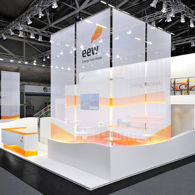 Ein Messestand von EEW (Energy from Waste) mit einer hohen, transparenten Struktur, die das Logo und den Slogan „Taking the lead.“ trägt. Der Stand hat geschwungene Wände und integrierte LED-Lichtbänder in Orange. Im Vordergrund befindet sich ein Empfangstresen mit Beleuchtung, während im Inneren Sitzbereiche mit weißen Tischen und Stühlen erkennbar sind. Eine erhöhte Plattform mit Glasgeländer im Hintergrund bietet zusätzliche Besprechungsmöglichkeiten. Die Szene wird durch eine helle, professionelle Beleuchtung ergänzt.