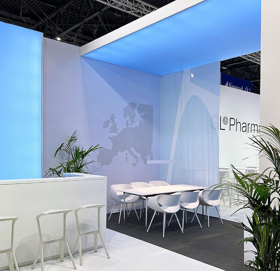 GL Pharma Messestand mit Besprechungsbereich, weißen Stühlen und Tisch. Blau beleuchtete Decke, Pflanzen und moderne, helle Gestaltung.