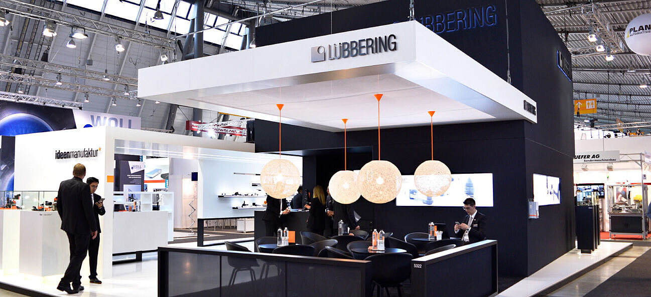 Messestand von LÜBBERING in Schwarz und Weiß mit hängenden runden Lampen und einer offenen Lounge mit schwarzen Sesseln und Tischen. Personen in Businesskleidung führen Gespräche, während im Hintergrund weitere Ausstellerstände sichtbar sind. Das Design wirkt hochwertig und modern.