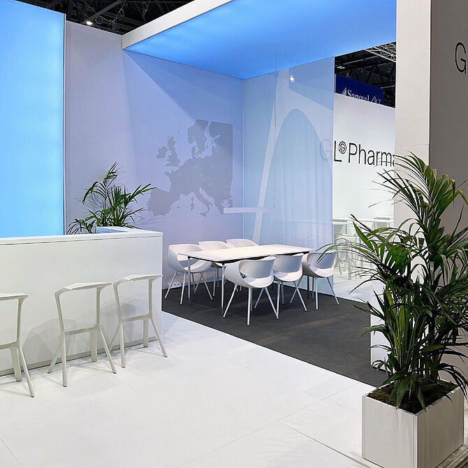 GL Pharma Messestand mit weißen Stühlen und Tischen, einem Tresen, dekorativen Pflanzen und einer stilisierten Europakarte an der Wand.