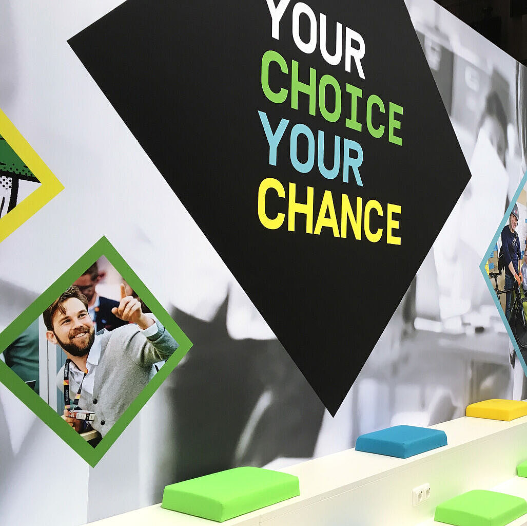 Weiße Tribüne mit bunten Sitzpolstern (grün, blau, gelb) vor einer Wand mit dem großen Schriftzug „YOUR CHOICE YOUR CHANCE“; daneben mehrere gerahmte Farbfotos von jungen Menschen in verschiedenen Szenen – lächelnd, diskutierend und mit Virtual-Reality-Brille.