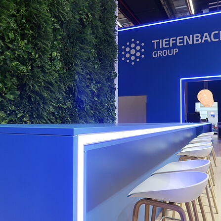 Nahaufnahme einer modernen blauen Theke mit integrierter LED-Beleuchtung. Davor stehen mehrere weiße Barhocker mit Holzbeinen. Links ist eine große vertikale Pflanzenwand mit dichten grünen Blättern zu sehen. Im Hintergrund leuchtet das Logo der „Tiefenbacher Group“ auf einer blauen Wand.