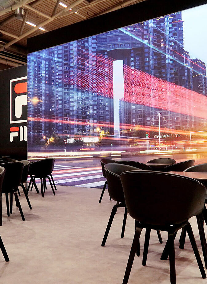 Ein FILA-Messestand mit einer stilvollen Lounge-Bereich, bestehend aus mehreren schwarzen Tischen und modernen Stühlen. Im Hintergrund ist eine große LED-Wand zu sehen, die eine belebte Stadtstraße mit Lichtspuren zeigt. Links daneben befindet sich eine FILA-Wand mit Logo sowie Regale mit Sneakers und ein Schaufensterpuppe in sportlicher Kleidung. Der Stand kombiniert eine urbane, moderne Ästhetik mit einer gemütlichen Atmosphäre.