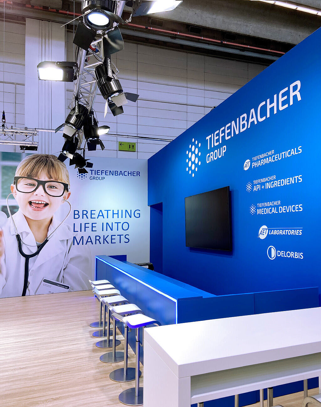 Messestand der Tiefenbacher Group mit einer blauen Theke und fünf weißen Barhockern. Im Hintergrund ein großes Banner mit einem lachenden Kind in Arztkittel und großer Brille, das ein Stethoskop um den Hals trägt. Daneben befindet sich eine blaue Wand mit Firmenlogos und einem Bildschirm. Über der Szene hängt eine Metallkonstruktion mit mehreren professionellen Strahlern. Der Boden ist aus hellem Holz.