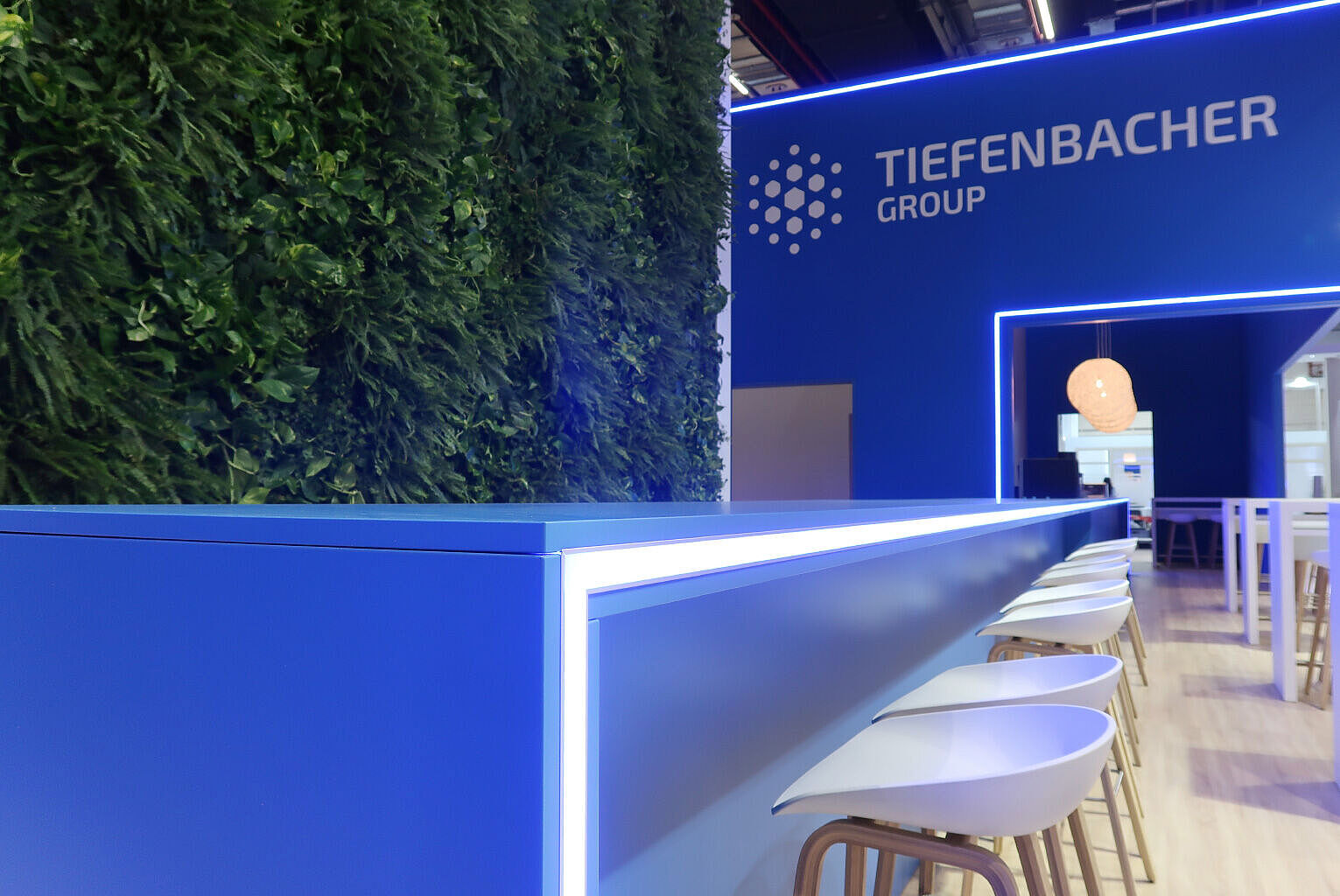 Moderner Messe-Stand der Tiefenbacher Group mit stilvollem Design in Blau, LED-Beleuchtung und einer grünen Pflanzenwand. Hochwertige Barhocker und Holz-Elemente schaffen eine einladende Atmosphäre.