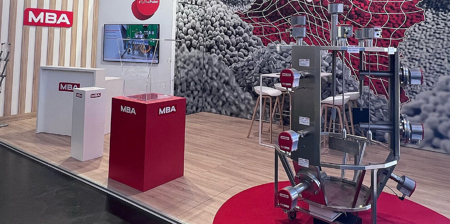 Ein Messestand mit Holzoptik-Boden zeigt mehrere Präsentationsflächen mit MBA-Branding, eine große technische Apparatur mit Metallrohren und Sensoren auf einem roten Teppich sowie eine auffällige Wandgrafik mit roten und grauen Pellets. Ein Bildschirm an der Wand zeigt eine digitale Präsentation.